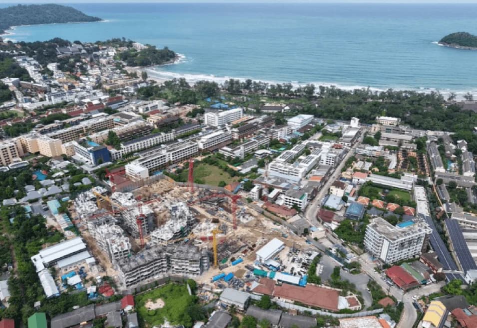 Construction, August 2026: the concrete frames of several wings with tower cranes, Kata Bay and Koh Pu behind